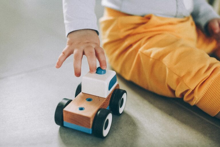 A baby playing with a wooden toy car on the floor wearing yellow pants.